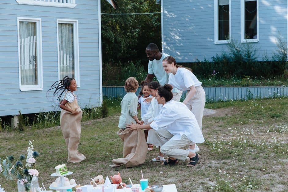 Joyful family gathering with children playing sack race in a garden outside a house.
